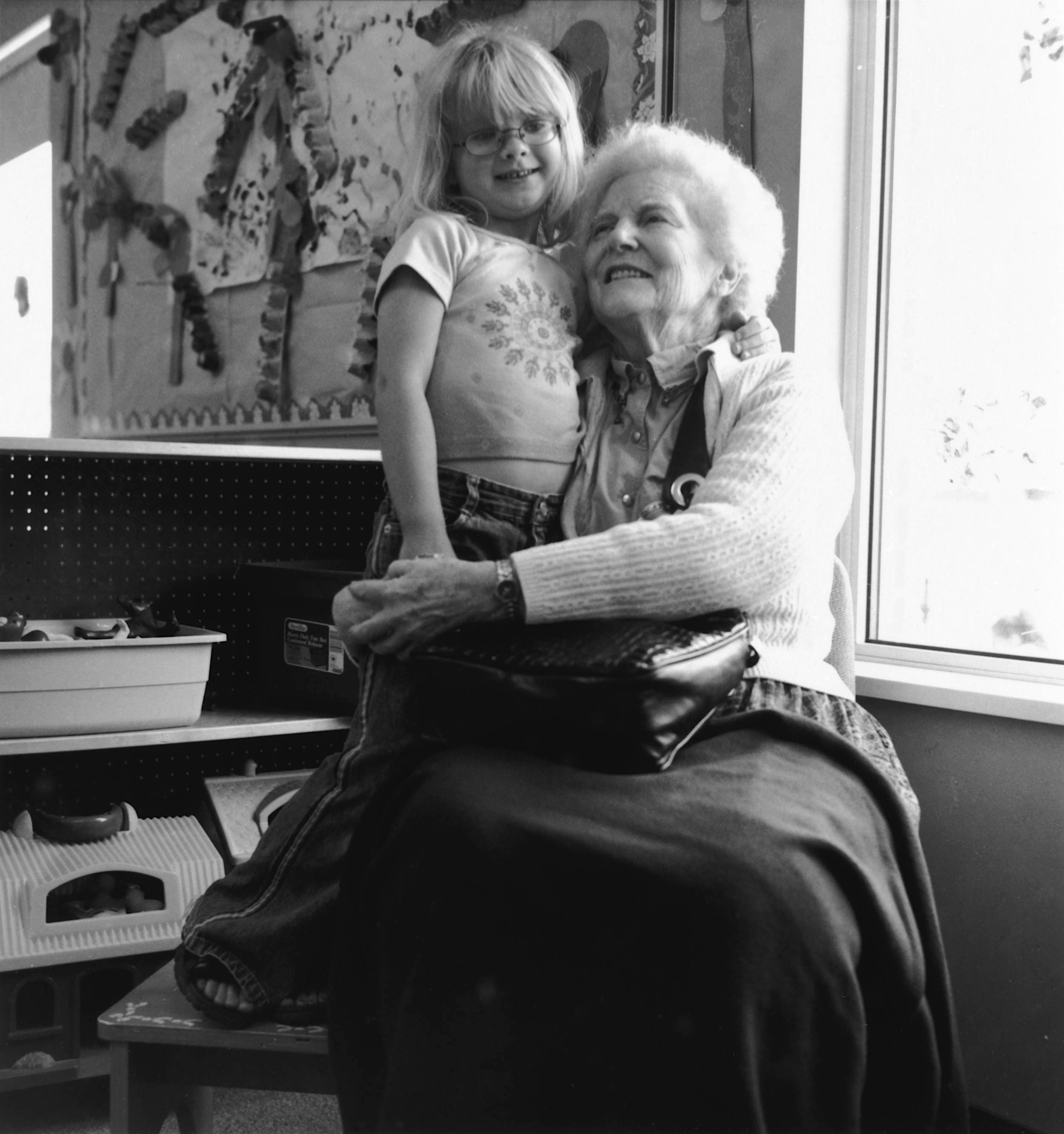 Black and white photo of elderly woman sitting down hugging young girl with both arms, both wearing warm smiles.