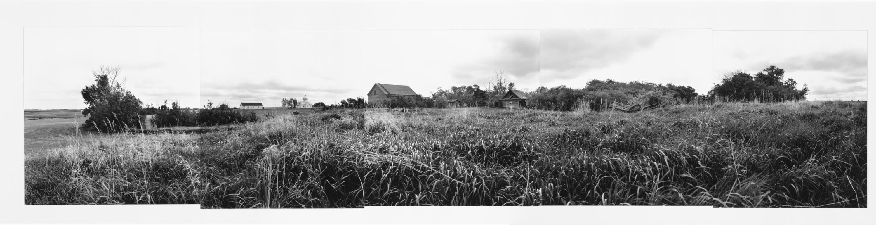 Black and white panorama of prairie landscape with some old buildings in distant background and tall grass in foreground.