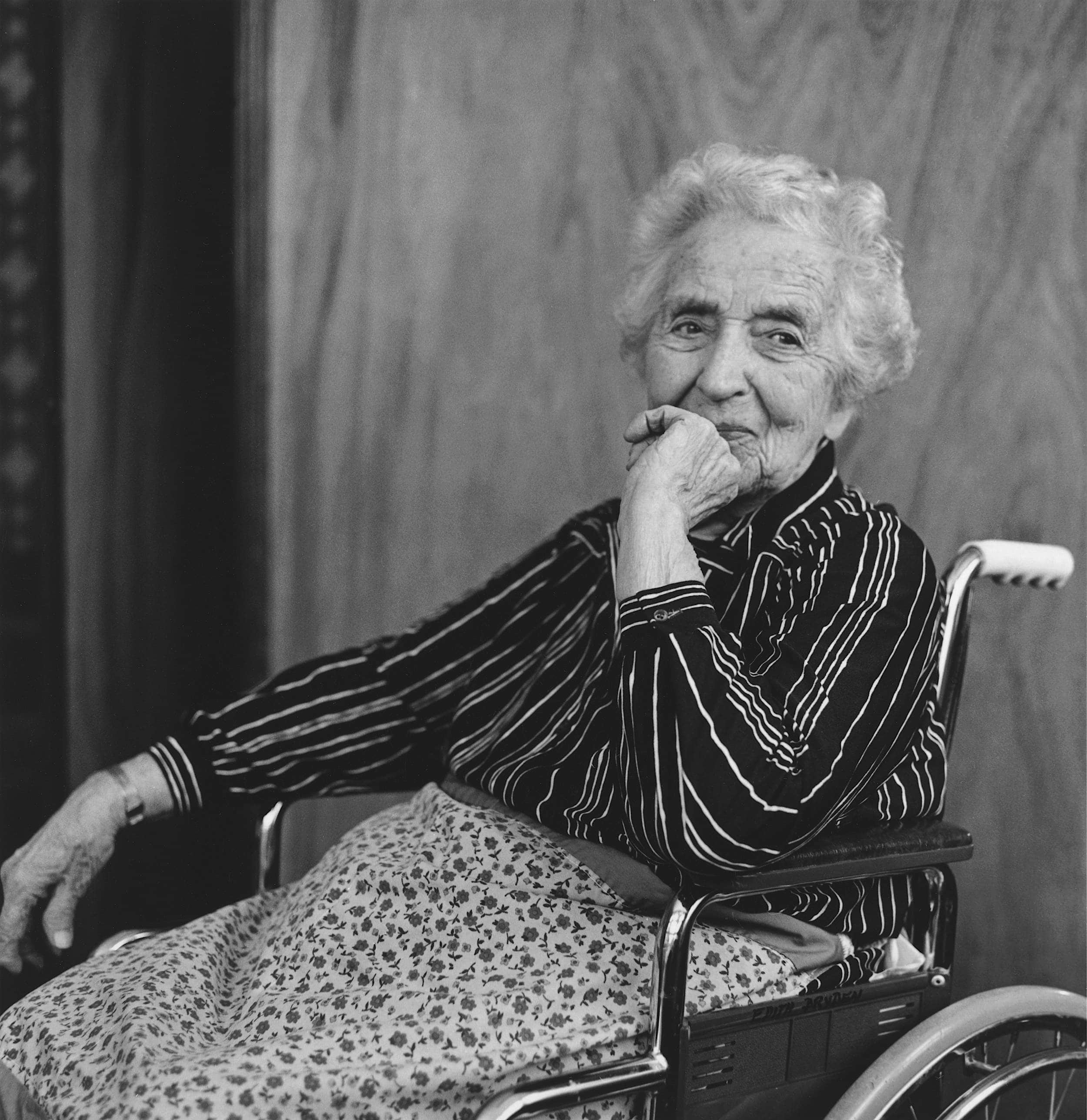 Black and white photo of elderly woman sitting in wheelchair on an angle with hand resting on chin.