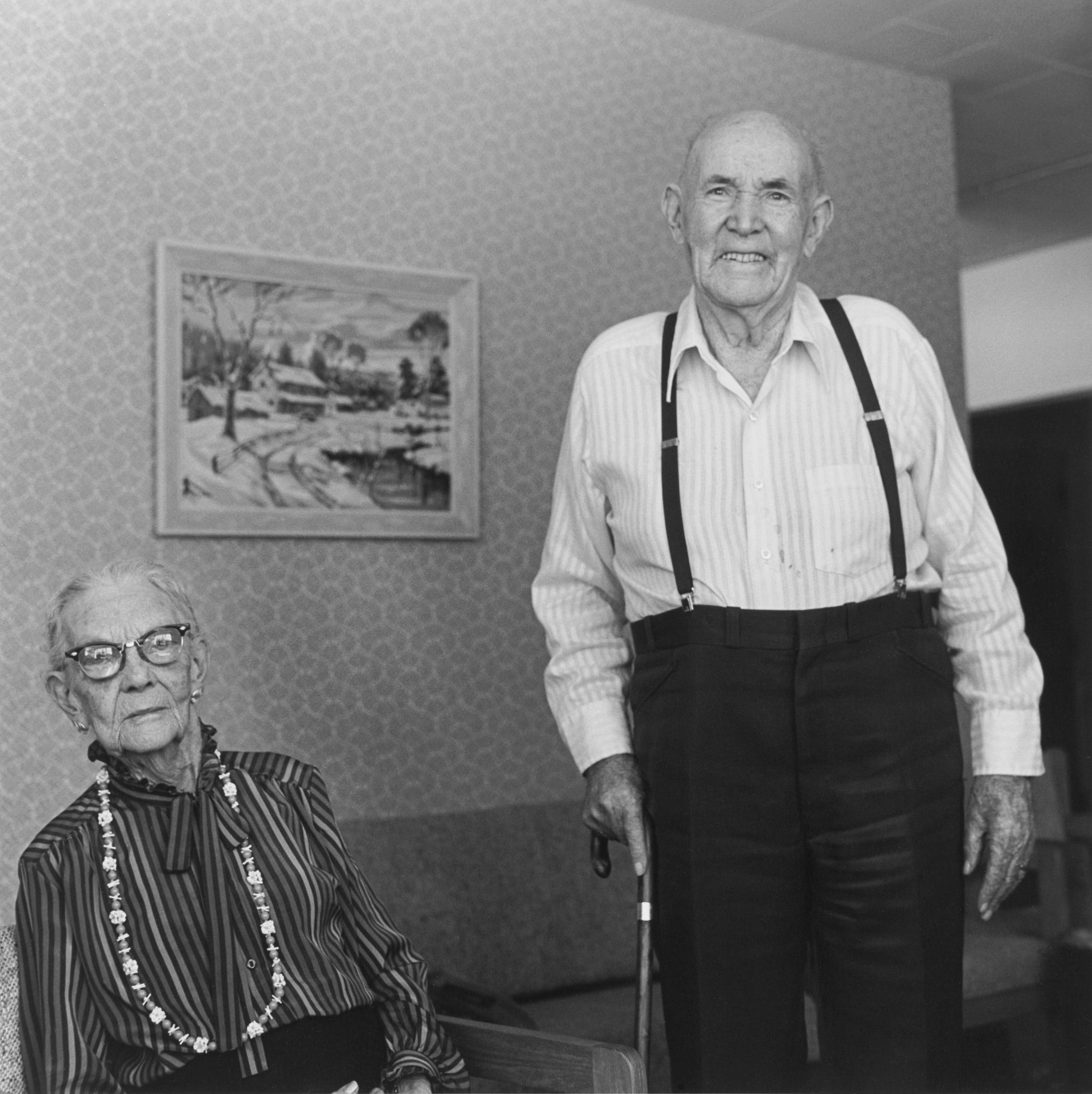Black and white photo of elderly couple, woman seated wearing beautiful large necklace and husband standing tall wearing suspenders, holding cane in right hand.
