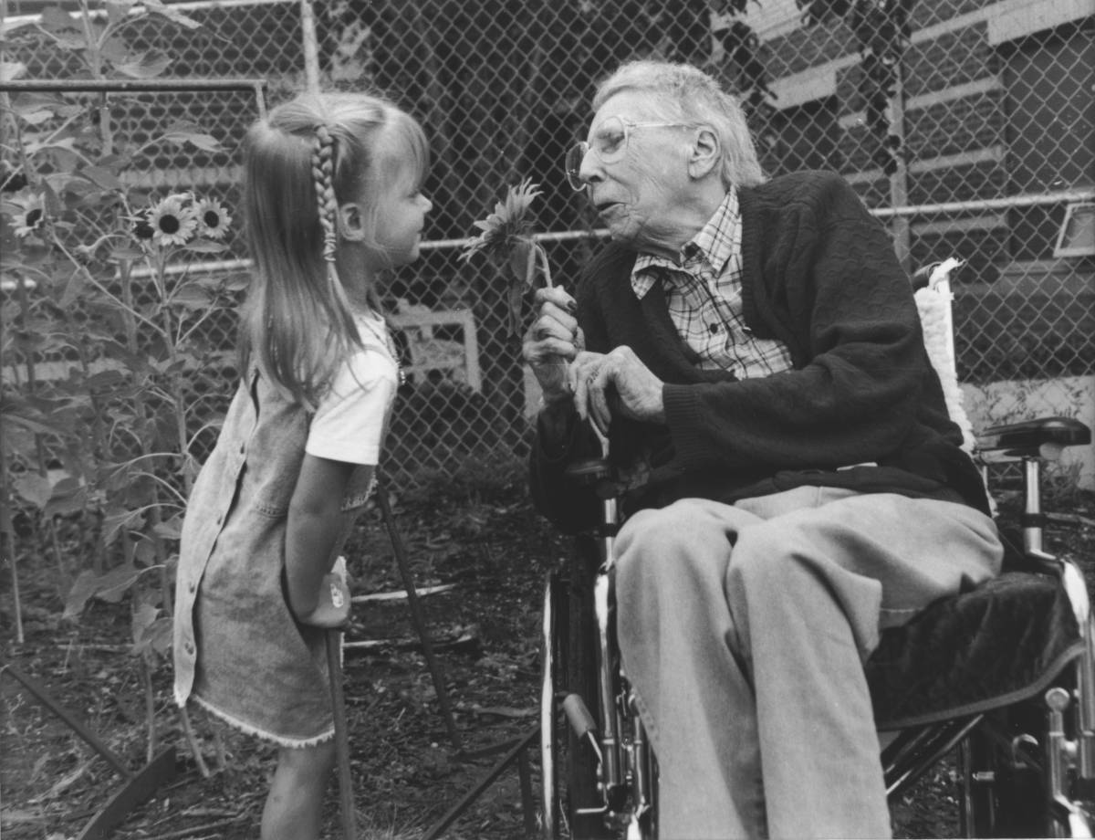 Black and white photo: elderly woman in wheel chair holder flower from young girl beside her.