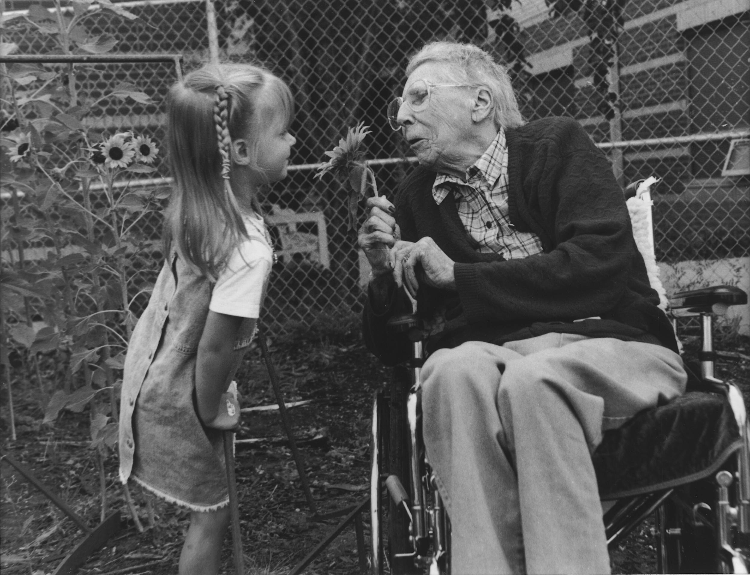 Black and white photo of elderly woman in wheelchair and young girl. Woman holding sunflower in hand as young girl looks at flower and woman affectionately.